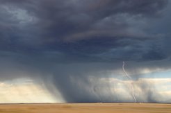 Das Bild zeigt eine dunkle Wolkenfront über Land, starken Regen und einen Blitz