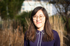 Das Bild zeigt eine junge Frau, Mengchen Dong, im Portrait mit Natur, Gräsern und einem Gebäude im unscharfen Hintergrund.
