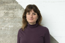 Das Bild zeigt eine junge Frau, Lou Haux, im Portrait vor einer Sichtbetonwand, die fast diagonal farblich geteilt, in dem der rechte obere teil weiß gestrichen ist.