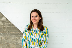The photo shows Ms. Elisa Buchberger in portrait in front of a concrete-like wall, diagonally divided and painted white in the upper right half.