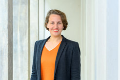 Kerstin Maria Pahl ist vom Bauch aufwärts vor einer weißen Betonwand zu sehen. Sie trägt ein orangefarbenes T-Shirt mit einem dunkelblauen Blazer darüber.