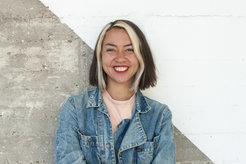 The photo shows Yayouk Willems (female) laughing in a portrait in front of a concrete-like wall, which is diagonally divided and painted white in the upper right half. She wears a light blue denim jacket and white strands in the front of her hair.