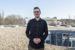 Nils Köbis (male) is standing on a roof covered with pebbles. He wears a dark shirt and clasps his hands in front of his stomach.