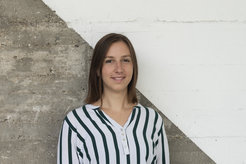 Das Foto zeigt Sonja Sudimac (weiblich) lächelnd im Portrait vor einer betonartigen Wand, die diagonal geteilt und in der oberen rechten Hälfte weiß gestrichen ist. Sie trägt eine weiß-schwarz gestreifte Bluse und hat offenes braunes schulterlanges Haar.