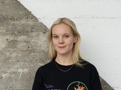 Das Foto zeigt Anika Löwe (weiblich) lächelnd im Portrait vor einer betonartigen Wand, die diagonal geteilt und in der oberen rechten Hälfte weiß gestrichen ist. Sie trägt ein schwarzes T-shirt mit Print und hat offenes blondes Haar.