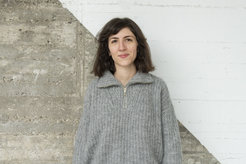 Das Foto zeigt Marwa El Zein (weiblich) lächelnd vor einer betonartigen Wand, die diagonal geteilt und in der oberen rechten Hälfte weiß gestrichen ist. Sie trägt einen grauen Pullover und hat offenes braunes Haar.