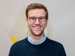 Das Bild zeigt Konstantin Offer (männlich) im Portrait vor einer weißen Wand direkt vor einem kleinen speziell designten Fenster. Er hat kurze Haare, trägt eine Brille und einen Pullover.