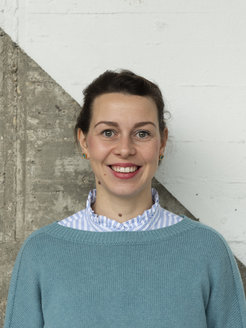 The photo shows Anne-Marie Nussberger (female) smiling in front of a concrete-like wall, which is divided diagonally and painted white in the upper right half. She is wearing a blue sweater over a blue and white striped blouse and has closed brown hair.