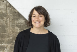 Das Foto zeigt Julia Wambach (weiblich) lächelnd vor einer betonartigen Wand, die diagonal geteilt und in der oberen rechten Hälfte weiß gestrichen ist. Sie trägt einen schwarzen Cardigan über einem schwarzen T-Shirt und hat kurzes, offenes, braunes Haar.