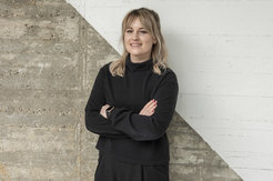 Das Foto zeigt Sarah Power (weiblich) lächelnd vor einer betonartigen Wand, die diagonal geteilt und in der oberen rechten Hälfte weiß gestrichen ist. Sie trägt einen schwarzen Pullover und hat offenes schulterlanges, blondes Haar.