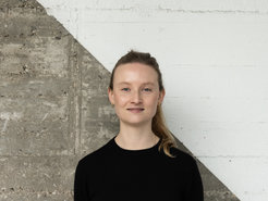 Das Foto zeigt Lisa Oswald (weiblich) lächelnd vor einer betonartigen Wand, die diagonal geteilt und in der oberen rechten Hälfte weiß gestrichen ist. Sie trägt einen schwarzen Pullover und hat ihr Haar zu einem Pferdeschwanz gebunden.