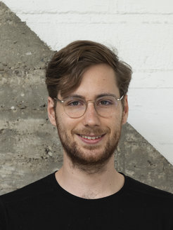 The picture shows Aaron Peikert (male) in front of a concrete wall separated by a diagonal gray and white line. He is wearing a black top. He has short brown hair and wears glasses and a beard.
