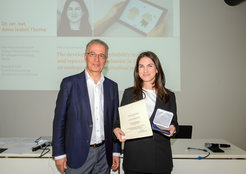 Im Bild steht Anna Thoma (weiblich) neben einem Mann im Anzug mit blauem Jackett. Sie hält erfreut ihre Urkunde in der linken und ihre gewonnene Medaille in der rechten Hand. Sie trägt ein weißes Shirt, darüber ein schwarzes Jackett.