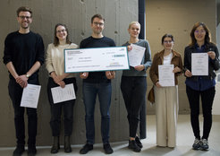 Die Teilnehmenden stehen mit ihren Urkunden nebeneinander vor einer Betonwand. In der Mitte steht der Gewinner des Awards Stefan Appelhoff (männlich) und präsentiert stolz seinen Scheck.