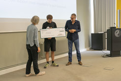 In the picture, Stefan Appelhoff, standing in the middle of the picture, receives his check and looks down at it. Maike Kleemeyer (female) is standing to his left and Ralph Hertwig (male) to his right. Both are also looking at the check.