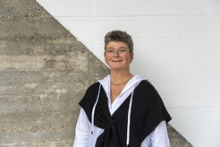 Das Foto zeigt Sabrina Strelow (weiblich) lächelnd vor einer betonartigen Wand, die diagonal geteilt und in der oberen rechten Hälfte weiß gestrichen ist. Sie trägt einen weißen Pullover mit einer schwarzen Weste darüber. Sie hat kurze blonde Haar und trägt eine Brille im Gesicht.