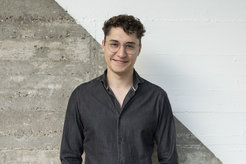 Das Foto zeigt Hannes Diemerling (männlich) lächelnd vor einer betonartigen Wand, die diagonal geteilt und in der oberen rechten Hälfte weiß gestrichen ist.Er trägt ein offenes, schwarzes Hemd. Er hat kurzes, braunes Haar und trägt eine Brille.