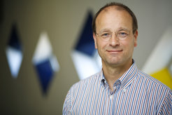 Männliche Person in blau-weiß gestreiftem Hemd steht vor unscharfem Hintergrund, der geometrische Formen in Blau und Weiß zeigt.
