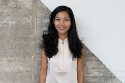 The photo shows Chi (Zoe) Ngo (female), smiling in front of a concrete-like wall that is divided diagonally and painted white in the upper right half. She is wearing a white top. She wears her long, black, curly hair loose.