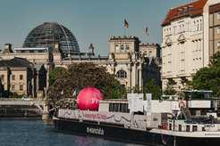 The iconic Reichstag dome in Berlin, adorned with German flags, overlooks a river where the ship "MS Wissenschaft" displays a banner for the "Wissenschaftsjahr 2024 Freiheit."