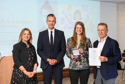 The picture shows Sina Schwarze (female) standing between two men in suits with blue jackets. On the far left, there is another smiling woman in a black dress. Sina is happily holding her medal up to the camera while the man to her right presents her with her certificate. Sina is wearing a dark blue jacket with flowers on it and black trousers. Her long blonde hair falls in soft waves over her shoulders.
