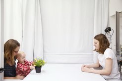 Eine Mutter mit ihrem Kleinkind auf dem Schoß, vor ihnen eine Pflanze, ihnen gegenüber eine wissenschaftliche Assistentin