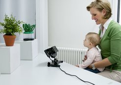 Dieses Bild zeigt eine Mutter, die mit ihrem Kind auf dem Schoß an einem Tisch sitzt. Das Kleinkind betrachtet zwei Objekte, die auf dem Tisch stehen. Bei dem Objekt links vom Kind handelt es sich um eine Topfpflanze und bei dem Objekt rechts vom Kind um ein von Menschen gemachtes Artefakt, das aus kleinen grünen Perlen besteht, die auf mehreren Drähten aufgezogen sind und auf einer durchsichtigen runden Box befestigt sind. Das Artefakt ähnelt einer Topfpflanze. Auf dem Tisch vor dem Kind befindet sich auch ein Eye-Tracker.